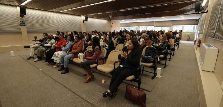Fotografia de uma plateia, formada por homens e mulheres, sentada em cadeiras pretas com estofad...