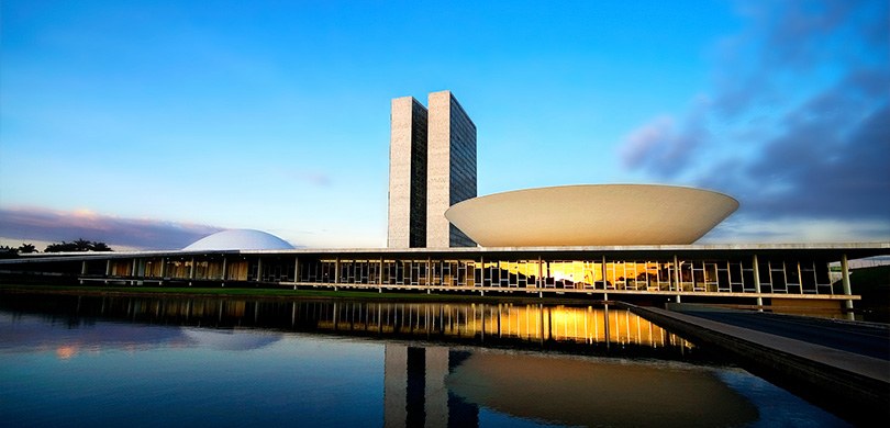 Prédios públicos onde estão instalados o Senado e a Câmara Federal fotografados ao entardecer. À...