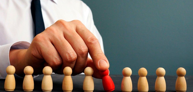 Série Assédio e Discriminação: saiba o que é e como denunciar discriminação no trabalho Na imagem, há vários bonecos de madeira sobre uma mesa cinza, uns atrás dos outros em uma fila h...