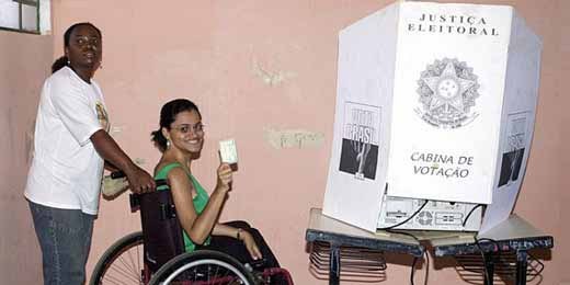 Fotografia de uma cadeirante com um ajudante em frente a uma cabine de votação
