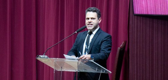 Fotografia de um homem falando em frente a um grupo de pessoas em um auditório. Ele usa uma blus...