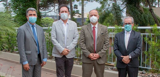 Servidor é homenageado pelos 32 anos de serviços prestados Fotografia de quatro homens em pé. Eles usam terno e máscara e olham para frente. Atrás, contorn...