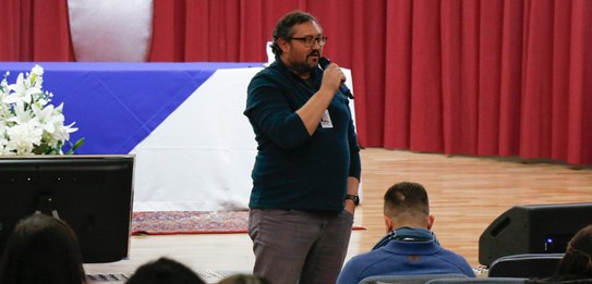 Fotografia de um homem falando em frente a um grupo de pessoas em um auditório. Ele usa uma blus...