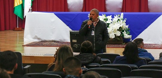 Fotografia de um homem falando em frente a um grupo de pessoas em um auditório. Ele usa uma blus...