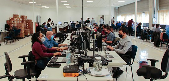 Fotografia de dois homens testando uma urna eletrônica, com outras urnas enfileiradas ao lado. A...