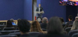 Fotografia de um auditório institucional durante uma apresentação ou palestra. Ao fundo, central...