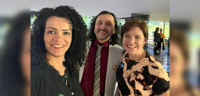 Fotografia em estilo selfie de três pessoas, duas mulheres e um homem, posando sorrindo. Todos v...