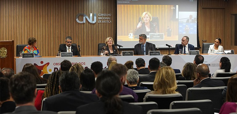 Fotografia de uma mesa de autoridades, com três mulheres e três homens, diante de uma plateia. A...