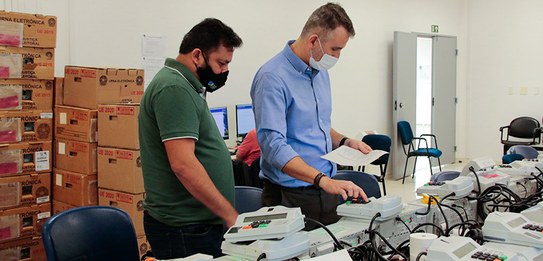Fotografia de dois homens testando uma urna eletrônica, com outras urnas enfileiradas ao lado. A...