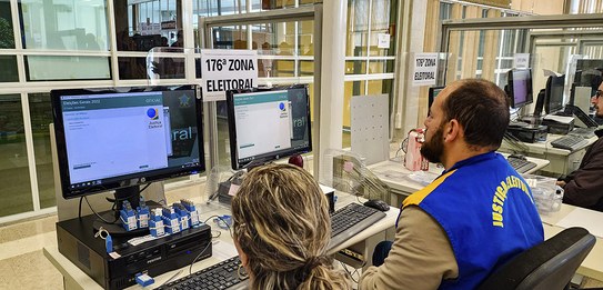 Fotografia de uma série de guichês de atendimento. Há um homem de terno e gravata em pé observan...