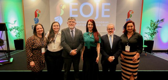 Fotografia de seis pessoas, quatro mulheres e dois homens, posando sorrindo em frente a um palc...