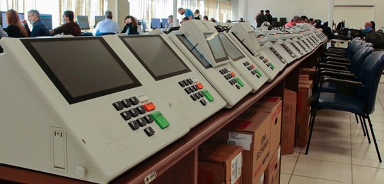 Fotografia de dois homens testando uma urna eletrônica, com outras urnas enfileiradas ao lado. A...