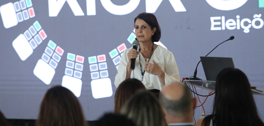 A Diretora-Geral Solange Maria Vieira fala para a plateia segurando um microfone. Atrás dela, um...