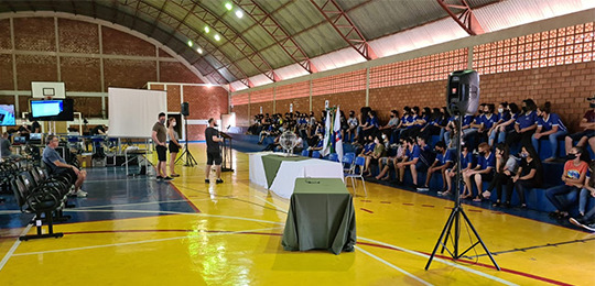 Foto em um ginásio de uma escola. À direita, há alunos sentados na arquibancada. No centro, há d...