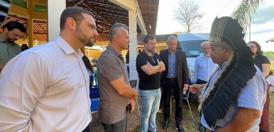 Fotografia em ambiente externo na qual se vê um grupo de homens em pé interagindo. À direita, em...