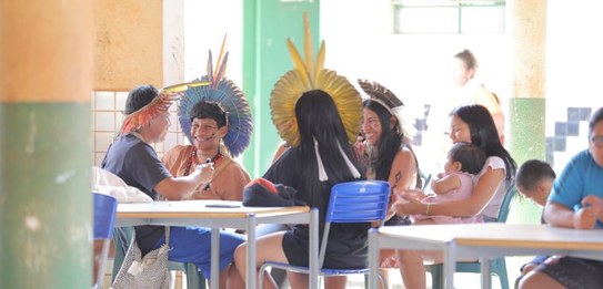 Fotografia em ambiente externo na qual se vê um grupo de homens em pé interagindo. À direita, em...