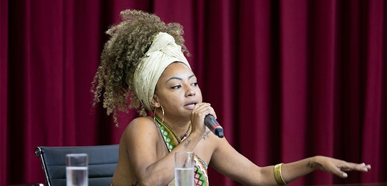 A fotografia mostra uma televisão e atrás um painel e uma cortina da cor vermelha. A tevê mostra...