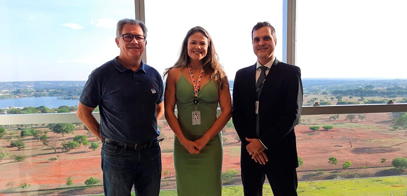 Fotografia de três pessoas estão em pé, lado a lado. Um homem usa camiseta polo e calça jeans, a...