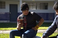 Um homem de gorro preto e camiseta escura com detalhes coloridos está sentado, bebendo de uma cu...
