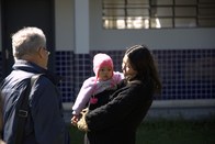 Uma mulher com cabelos escuros e uma jaqueta preta segura um bebê que usa um gorro rosa. Ela est...