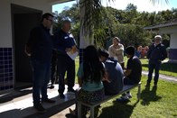 Um grupo de seis pessoas está reunido ao ar livre. Quatro pessoas estão sentadas em um banco e d...