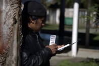 Um homem com boné e uma jaqueta de couro preta está encostado em uma palmeira. Ele segura um peq...