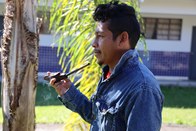 Um homem de jaqueta jeans está encostado em uma palmeira. Ele segura e sopra fumaça de um cachim...