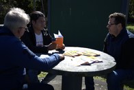 Dois homens estão sentados em uma mesa redonda ao ar livre, em um gramado. O homem de cabelos gr...