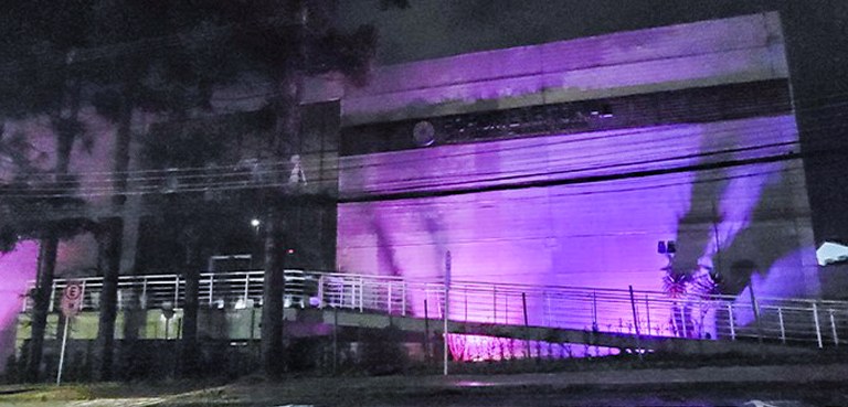 A foto mostra a fachada do Fórum Eleitoral de noite. Há luzes roxas clareando a frente do prédio...