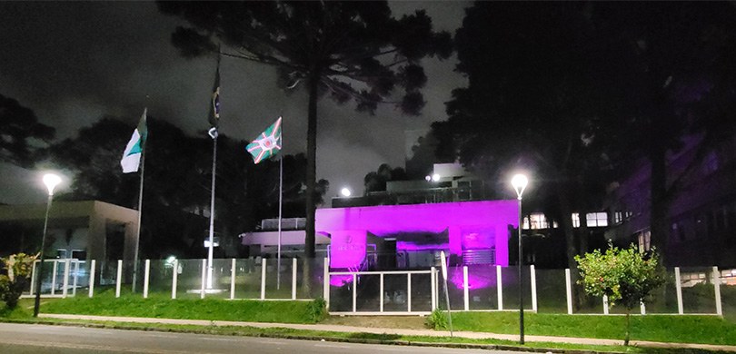 Fotografia noturna da fachada de um edifício iluminado com a cor roxa. Em primeiro plano, podem-...