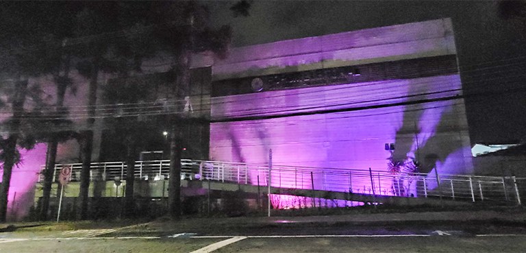 Fotografia da fachada do Fórum Eleitoral de Curitiba à noite iluminada na cor roxa. Na frente do...