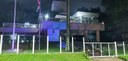 Fotografia do edifício-sede do TRE-PR iluminado na cor azul durante a noite. Ao redor, há alguma...