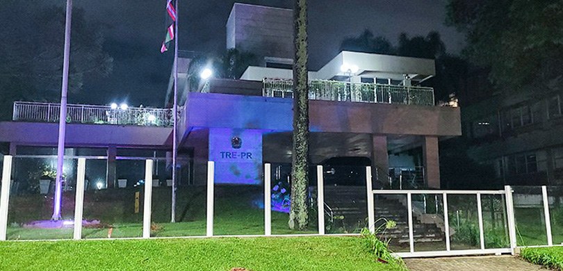 Fotografia do edifício-sede do TRE-PR iluminado na cor azul durante a noite. Ao redor, há alguma...