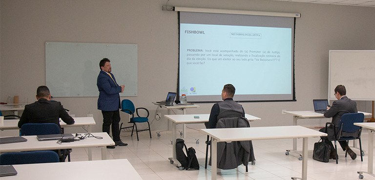 TRE-PR apresenta módulo de “Direito Eleitoral” no Curso Oficial de Formação Inicial do TJPR para juízas e juízes recém-empossados Fotografia do de um homem em pé ao lado de um telão diante de três homens sentados em carteiras ...