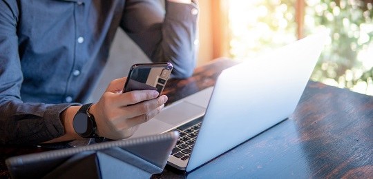 uma pessoa segurando um smartphone. uma mesa com um notebook aberto à frente.