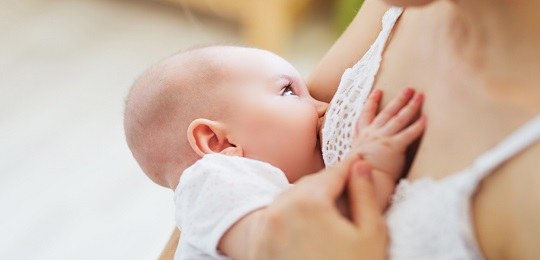 Fotografia de um bebê carequinha, vestindo body branco, mamando no peito da mãe, de quem se vê a...