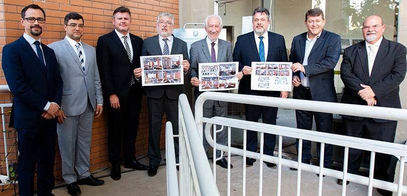 Foto em que oito homens vestindo ternos posam para a foto em frente a uma edificação. Do lado es...