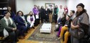 Fotografia de 17 mulheres reunidas em uma sala. Algumas aparecem sentadas e outras em pé, ao red...