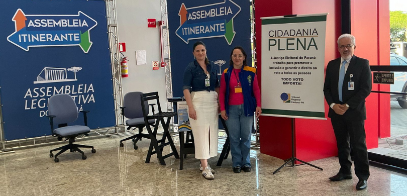 Duas mulheres e um homem posam para a foto em frente a dois banners azuis onde se lê Assembleia ...