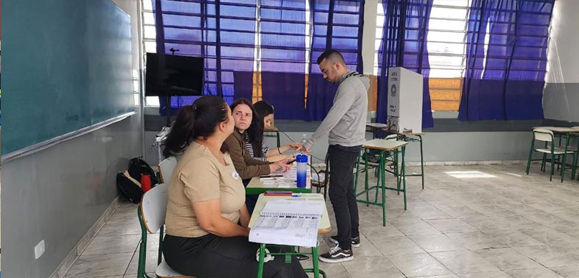 Fotografia de uma sala de aula. Um jovem está em pé diante de três mesárias, sentadas em carteir...