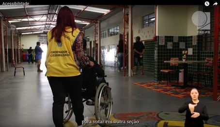 Foto do Coordenador de acessibilidade ajudando um cadeirante no local de votação. Algumas pessoa...