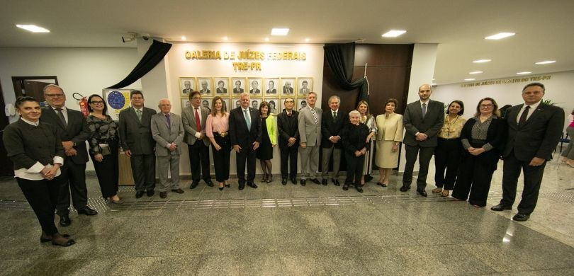 Várias mulheres e homens posam perfilados em frente a uma parede branca onde se lê: Galeria de J...