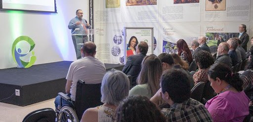 Fotografia de um homem segurando um microfone em cima de um palco, falando diante de uma plateia.