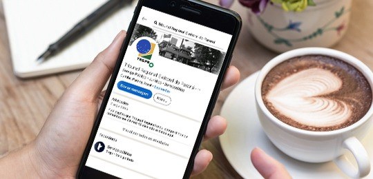 Imagem de uma mão segurando um celular com o perfil do TRE-PR no LinkedIn aberto. Ao fundo, uma ...