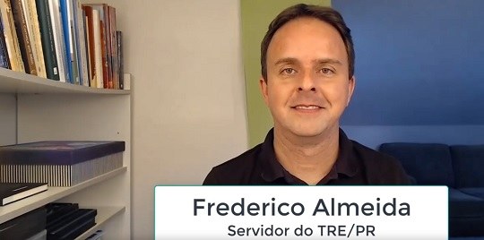 Fotografia de um homem vestindo uma camiseta preta. À esquerda há uma estante com livros