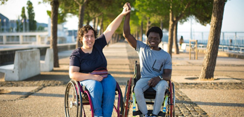 Fotografia de uma mulher e de um homem em uma cadeira de rodas. Eles estão sorrindo com os braço...