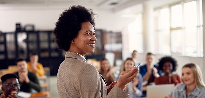TRE-PR parabeniza educadores pelo Dia dos Professores Fotografia de uma mulher diante de estudantes sentados em carteiras escolares. Em primeiro plano...