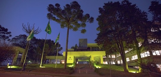 TRE-PR participa da campanha Setembro Amarelo Fotografia da fachada de um prédio iluminado com a cor amarela. Há uma Araucária e as bandeiras ...
