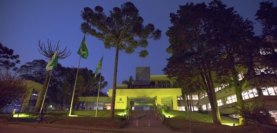 Fotografia da fachada de um prédio iluminado com a cor amarela. Há uma Araucária e as bandeiras ...