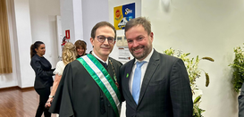 Foto de dois homens de terno posando juntos. O primeiro veste ainda uma toga e uma faixa verde e...
